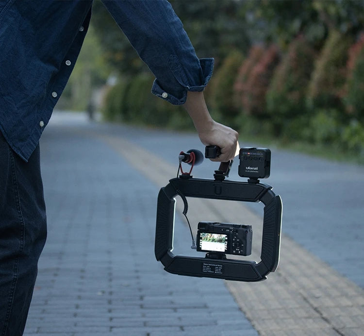 A person using a camera rig equipped with a digital camera, microphone, and an action camera, capturing video footage outdoors on a paved path.